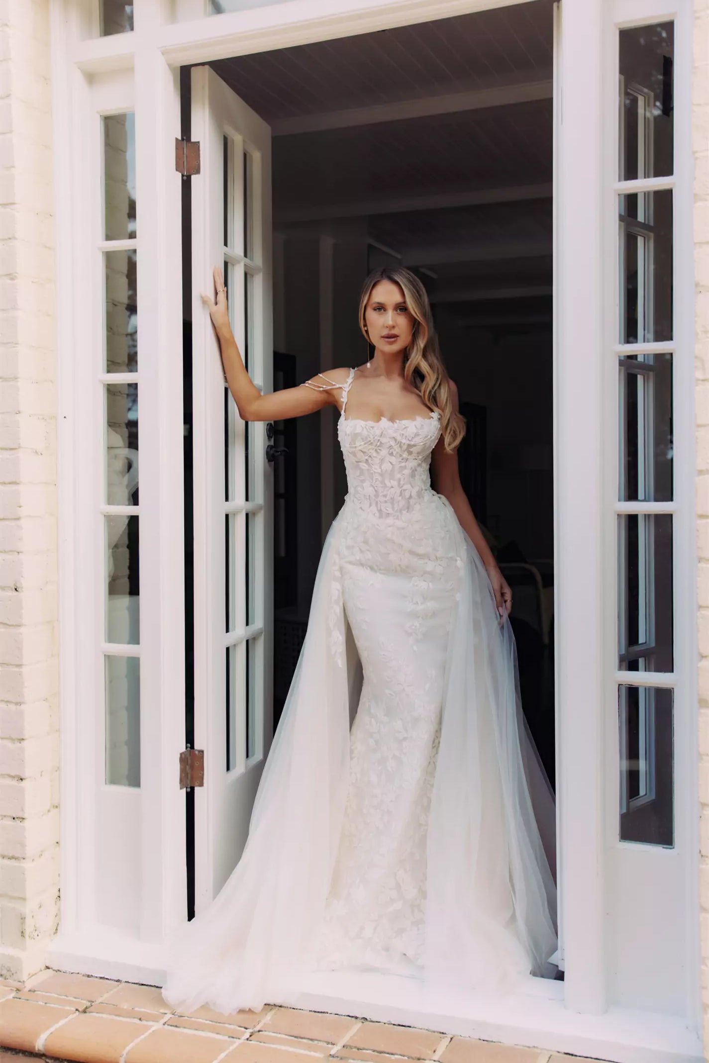 Bride in fitted lace wedding gown with illusion neckline and flowing tulle overskirt standing in white doorway — You&I Bridal | Endless UAE