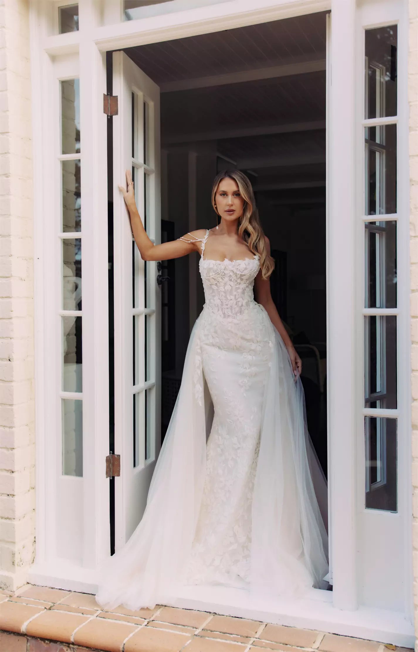 Bride in fitted lace wedding gown with illusion neckline and flowing tulle overskirt standing in white doorway — You&I Bridal | Endless UAE