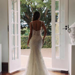 Bride in elegant lace mermaid wedding gown with open back and train, standing at French doors — You&I Bridal | Endless UAE
