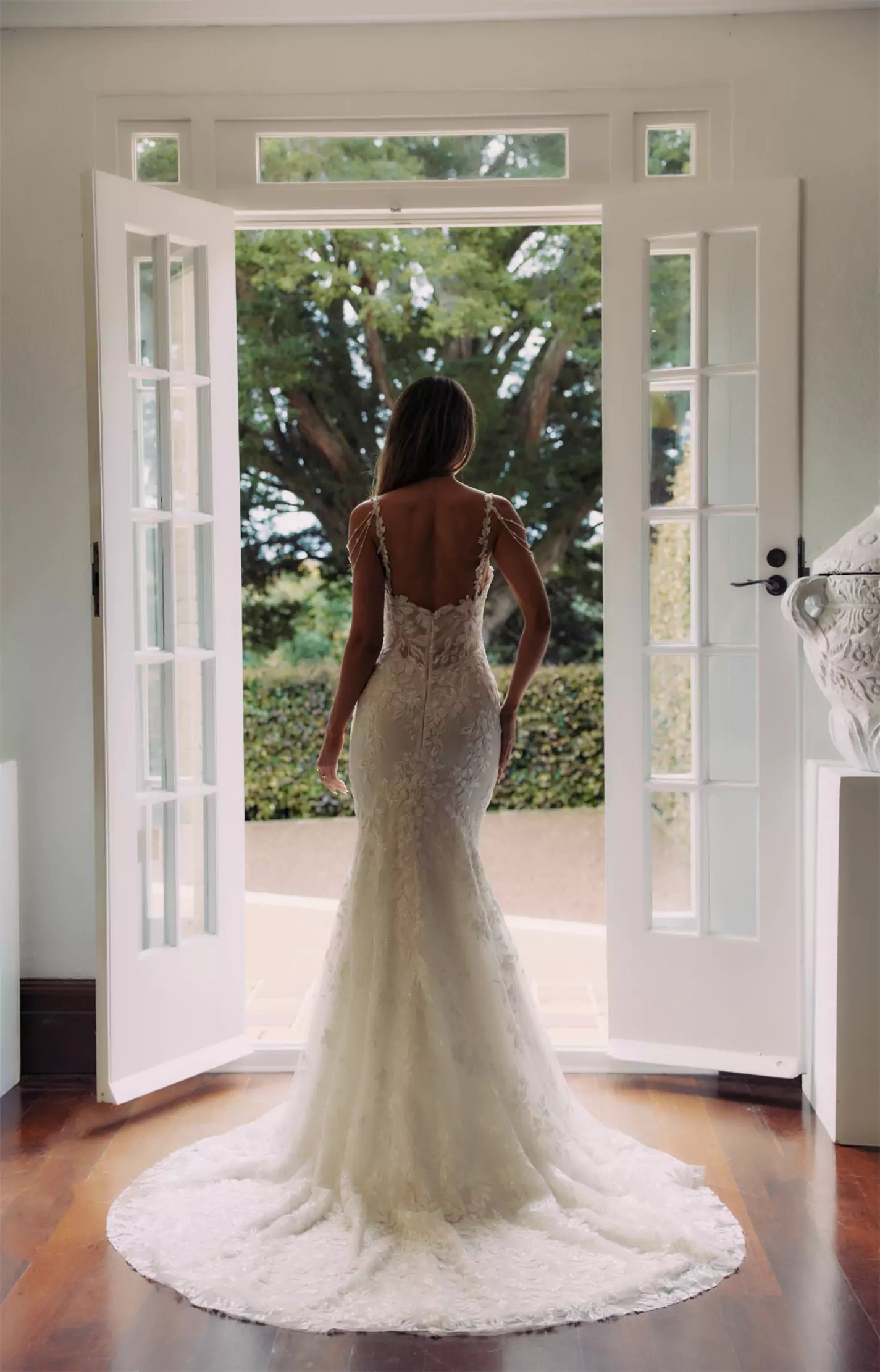 Bride in elegant lace mermaid wedding gown with open back and train, standing at French doors — You&I Bridal | Endless UAE