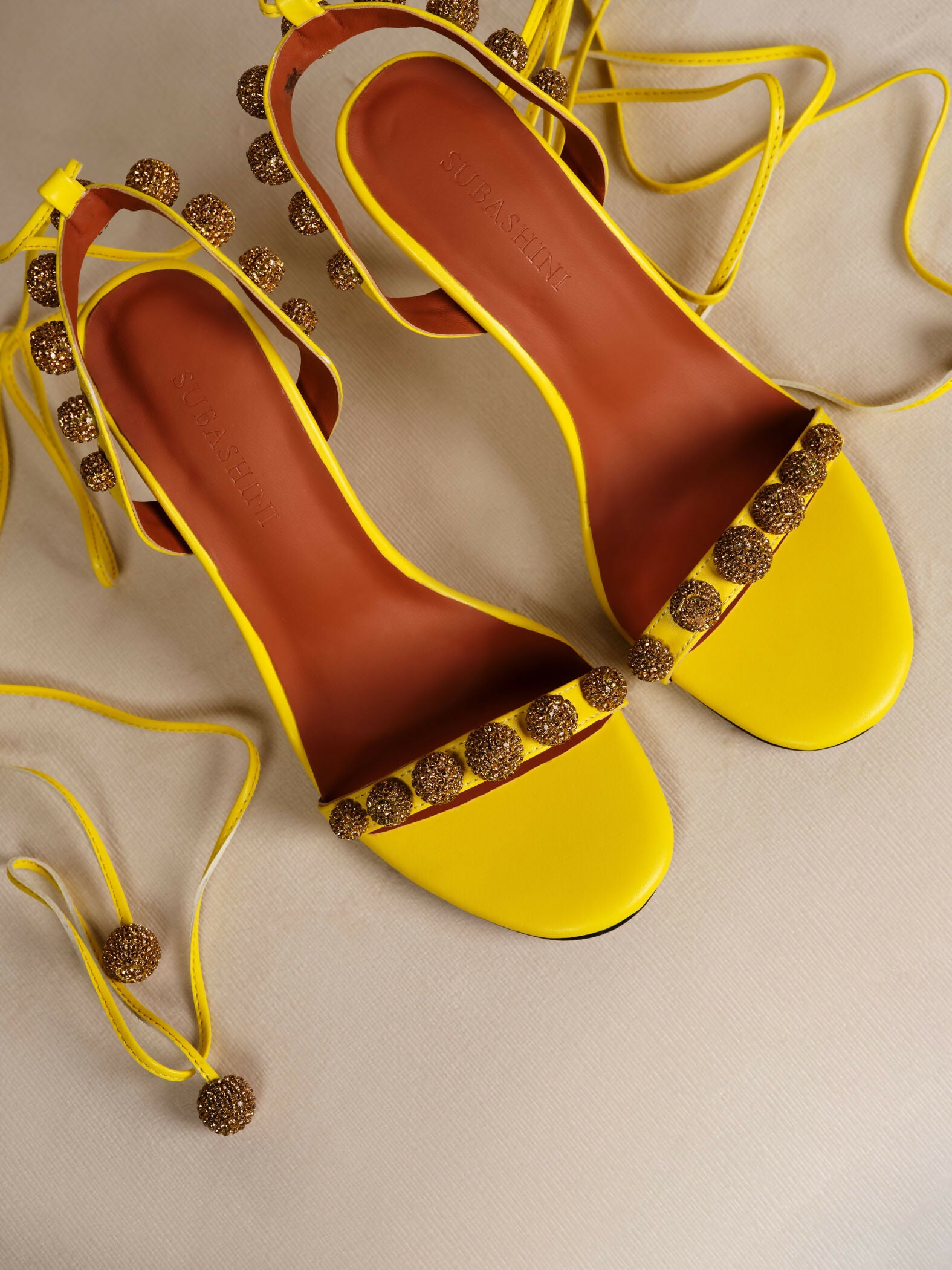 Bright yellow Bellaria heels with glittery gold embellishments and ankle-tie straps on beige background — Subashini Heels | Endless UAE