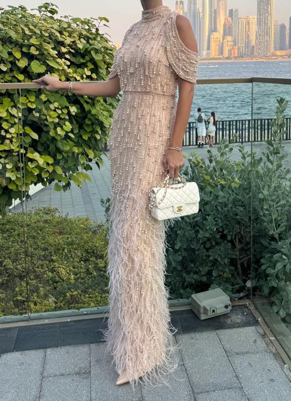 Champagne beaded gown with feather trim and cold-shoulder sleeves, paired with white quilted handbag — Pamella Roland | Endless UAE
