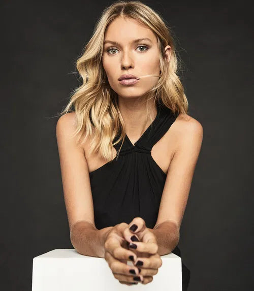 Blonde woman wearing black Hilda halterneck dress with twisted neckline detail, seated pose against dark background — Reiss | Endless UAE