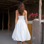 Woman in white strapless midi dress with pleated skirt and white heels against rustic wall — Cleopatress | Endless UAE