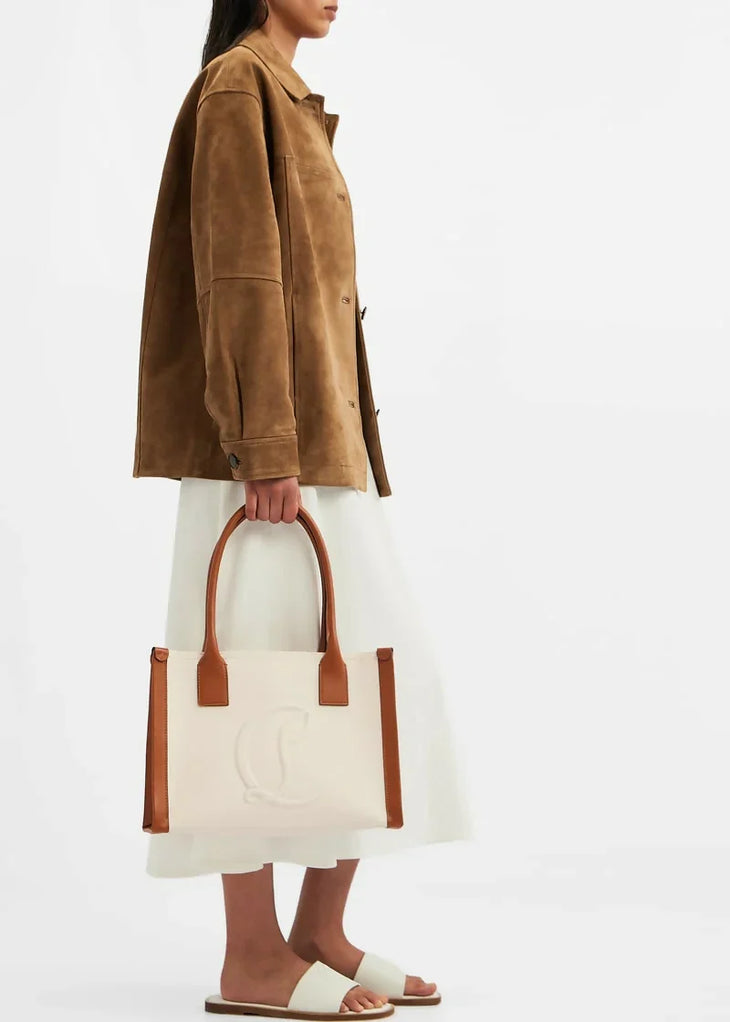 Woman holding cream canvas tote bag with brown leather handles and trim, wearing tan suede jacket and white skirt — Christian Louboutin | Endless UAE